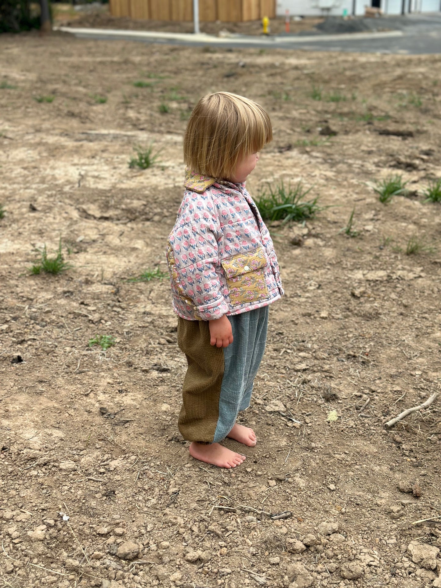 Child standing on a bare ground wearing a patterned jacket and jeans.