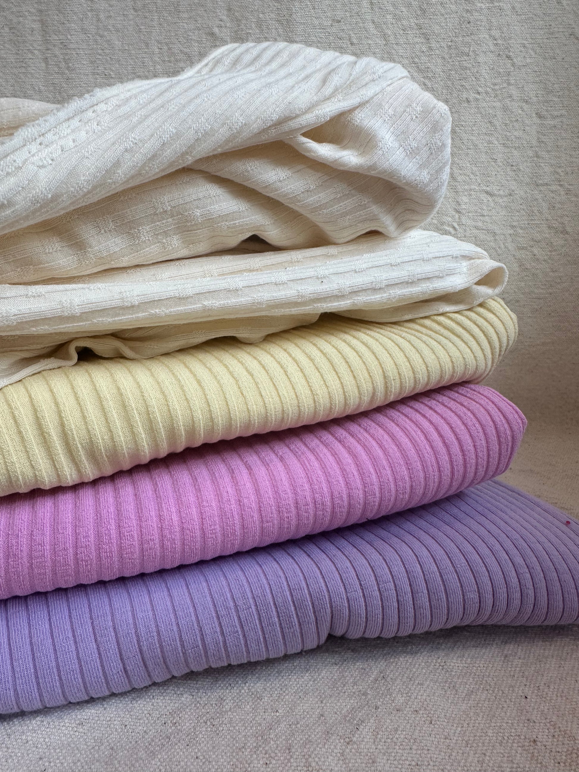 Stack of pastel rib knit fabrics in lilac, pink, buttercream, and natural ivory tones — soft 6x3 cotton spandex rib knit perfect for loungewear and cuffs.