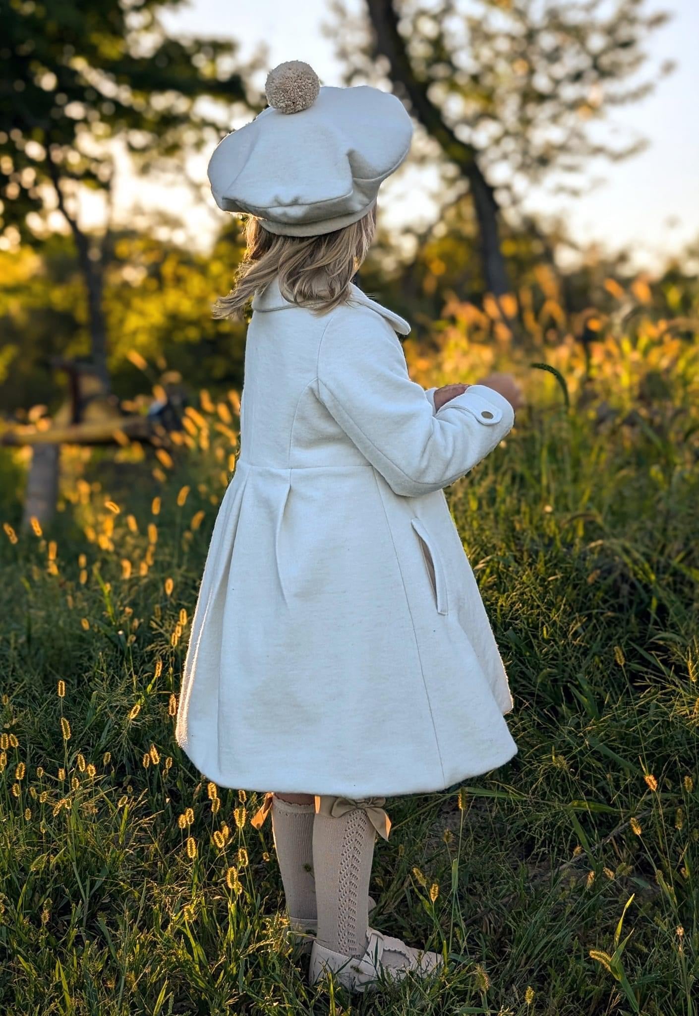 Girl in white coat and hat standing in tall grass.