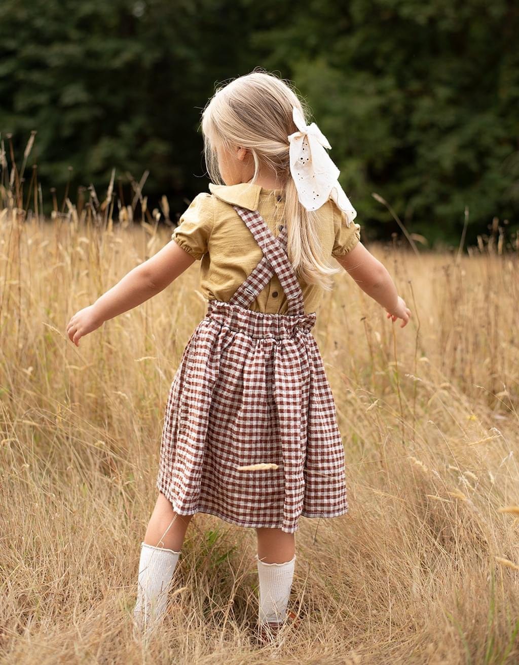 Blonde girl in plaid dress stands in a field.