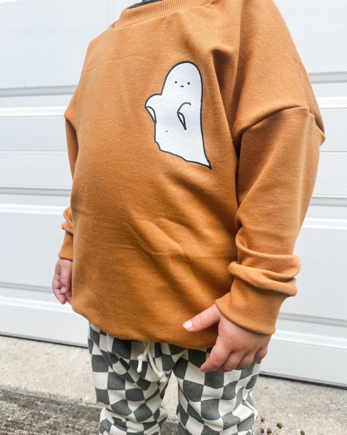 Child wearing a brown ghost sweatshirt and checkered pants.