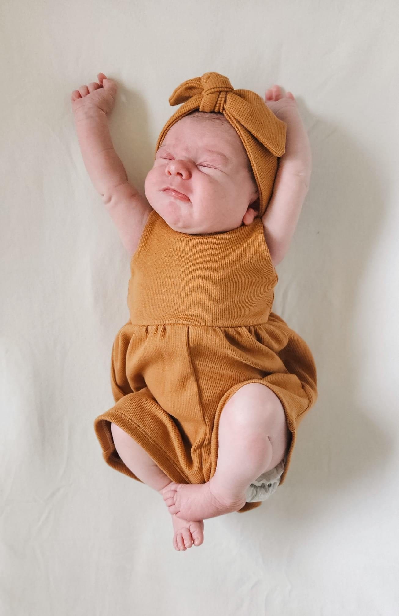 Baby in mustard-colored outfit and headband, arms raised.