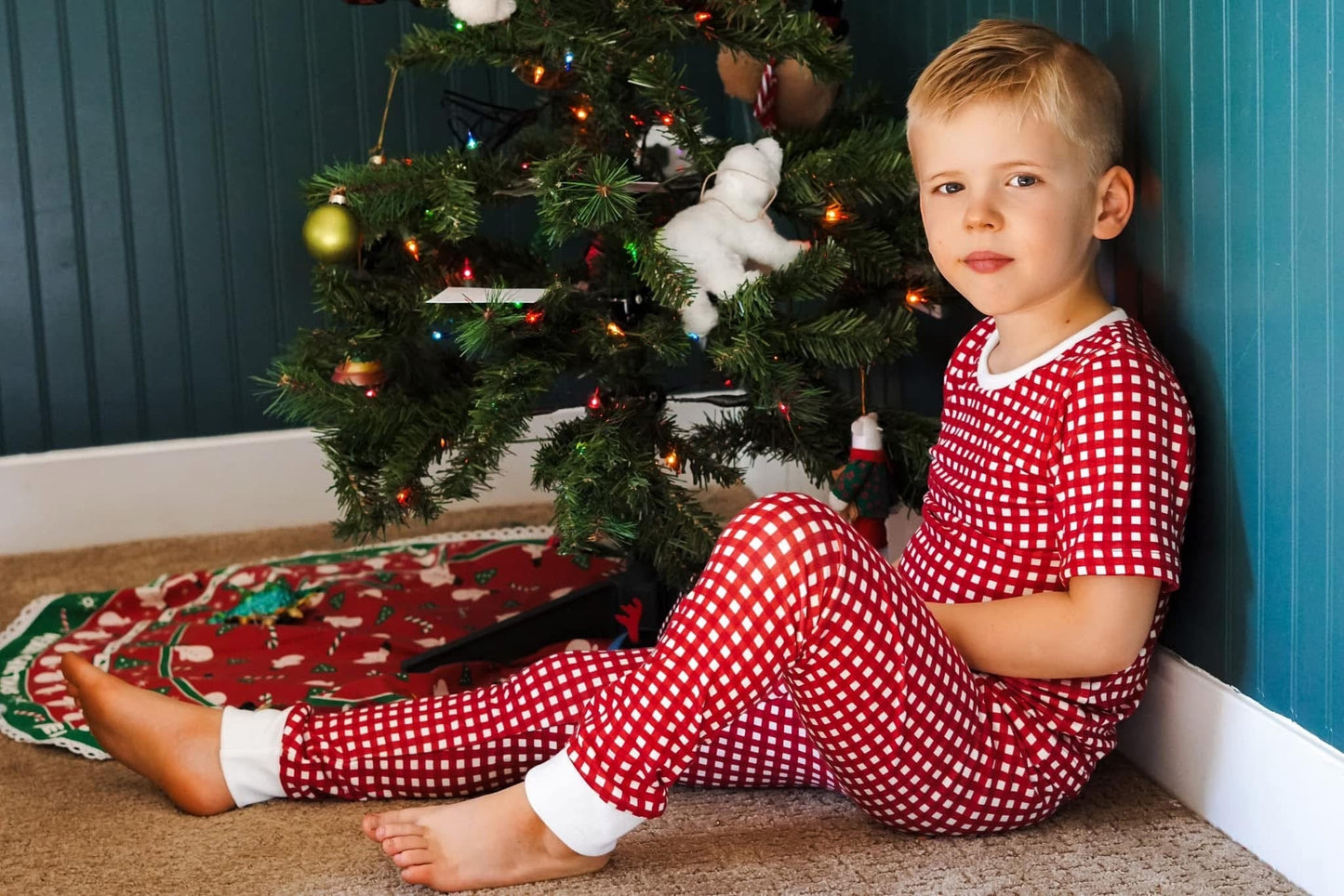 Boy in red pajamas sits by Christmas tree.