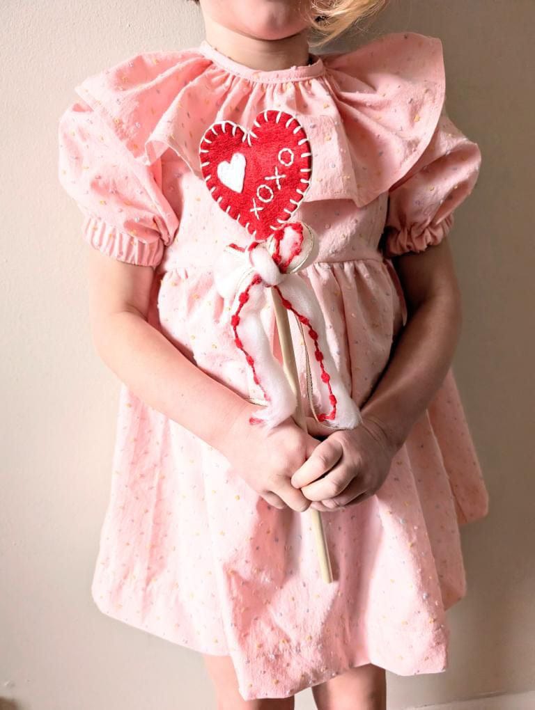 Child wearing pink dress holding heart-shaped lollipop.