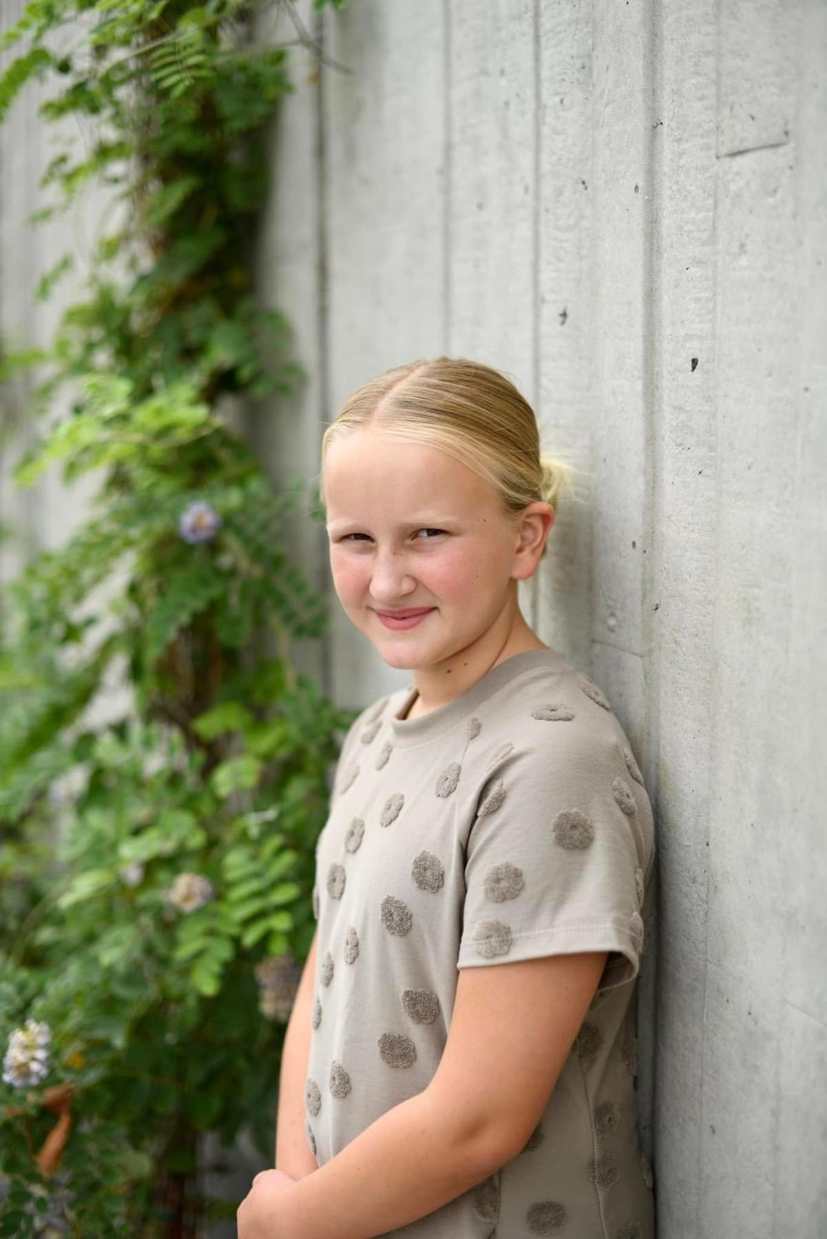 Blonde girl in tan shirt by a concrete wall and greenery.