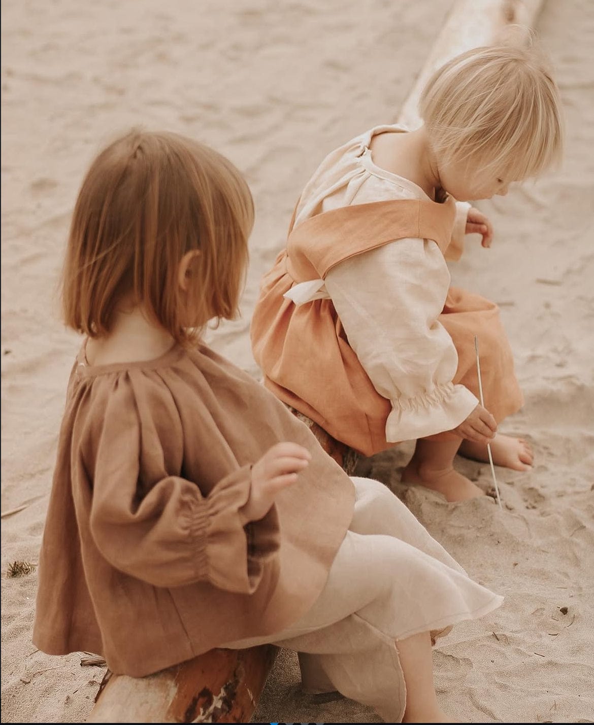 Two children in linen outfits sitting on sand.