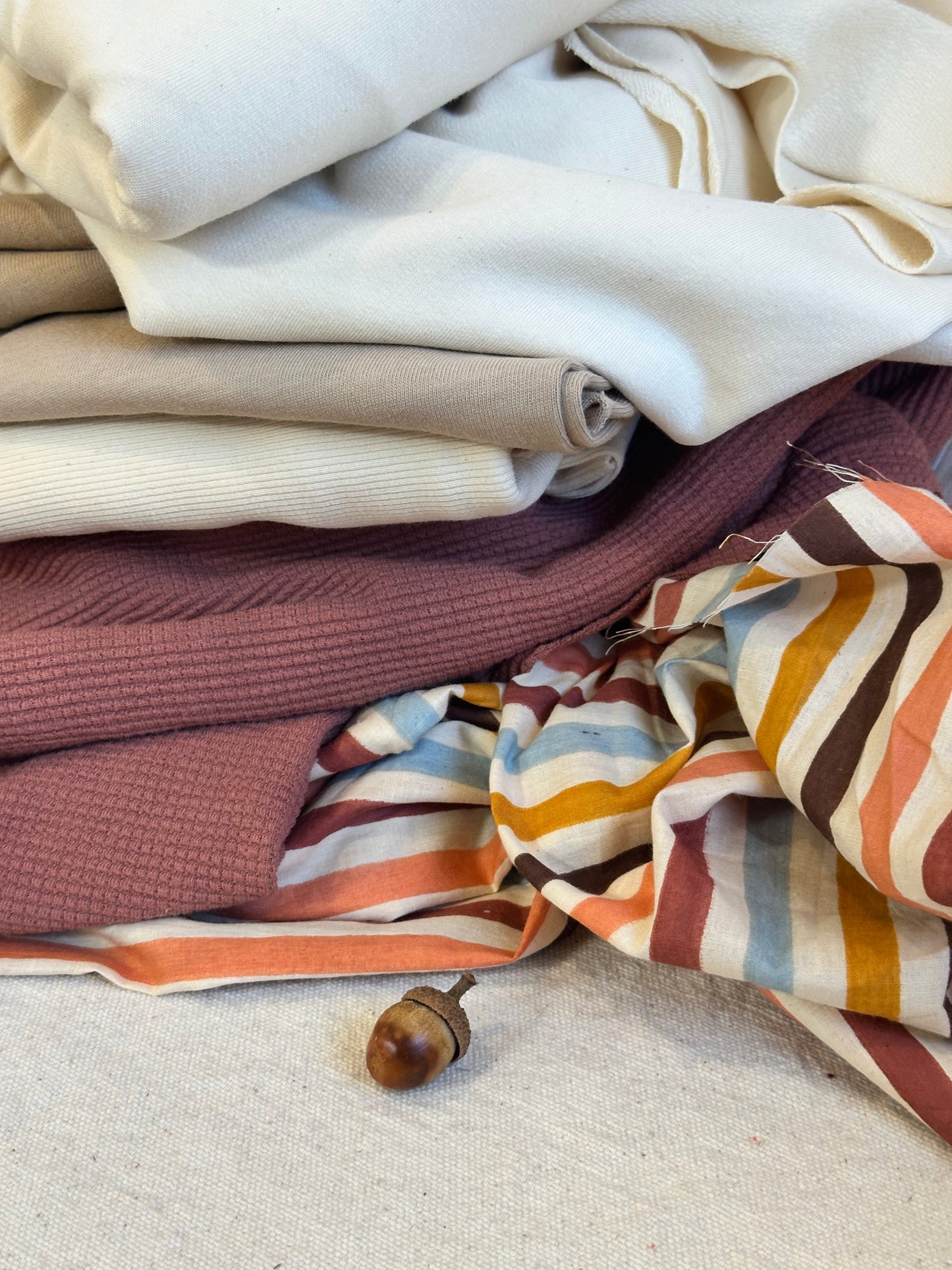 A styled flat-lay arrangement of folded and draped fabrics in neutral and striped colors, including cream, beige, dusty rose, and a striped textile with orange, brown, blue, and cream bands, with a small wooden acorn prop on a light fabric surface.